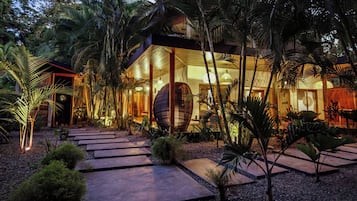 Uma casa moderna com uma grande fachada de vidro, cercada por vegetação tropical e um caminho de pedra.