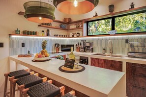 Cozinha moderna com área de bar, destacando uma bancada grande com uma tigela de frutas, uma taça de vinho e um letreiro de boas-vindas.