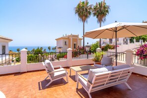 A terrace with white furniture, a table, and an umbrella. There are palm trees and a view of the sea. - Villa in Tamango Hill Near Playa Guilche (Nerja)