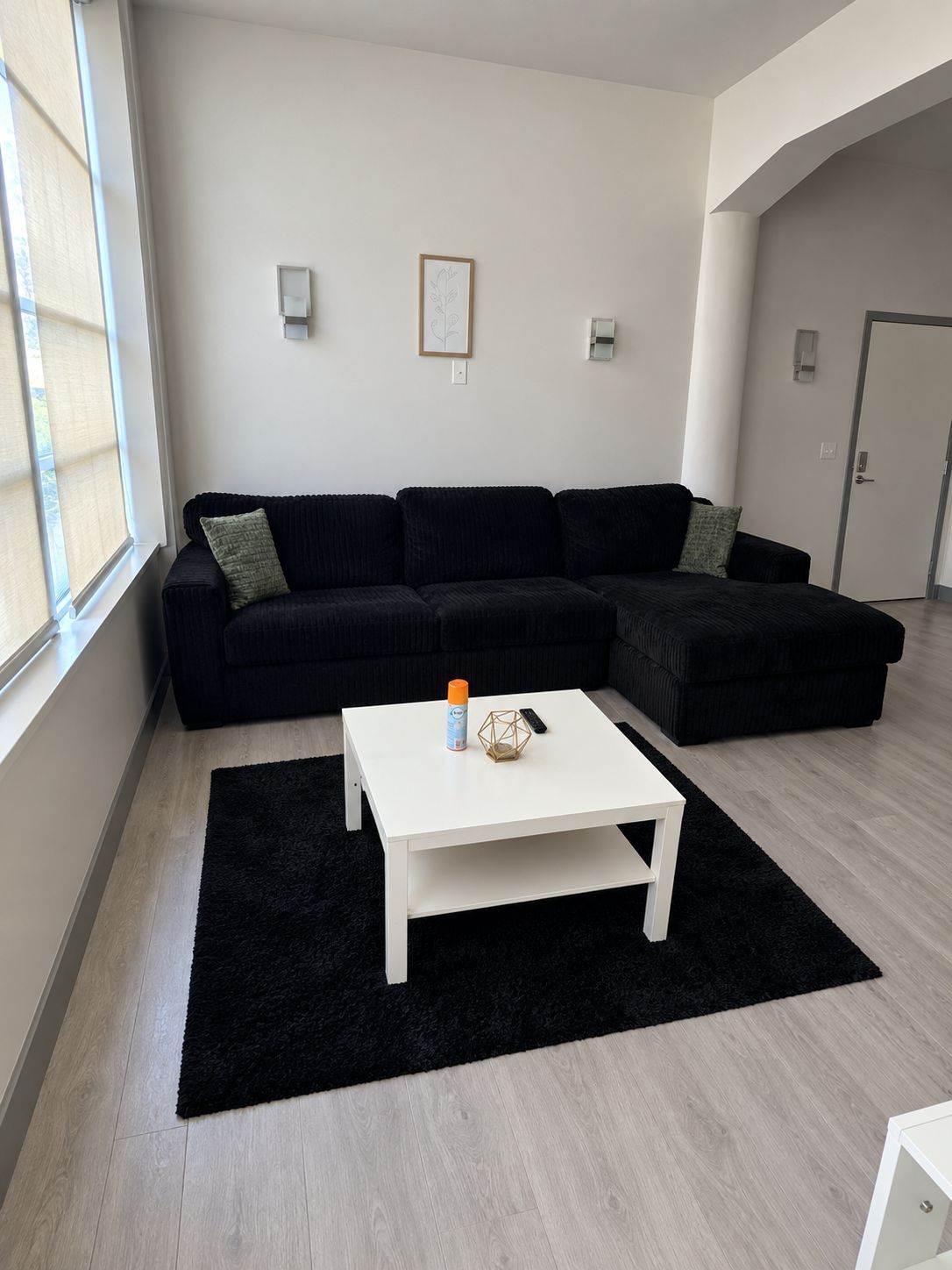 A modern living room with a black sectional sofa, a white coffee table, and a black rug. There are two wall-mounted lights and a framed picture on the wall.