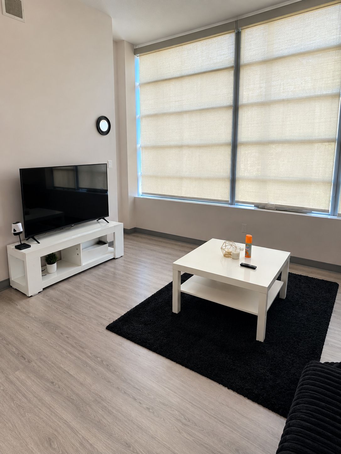 Uma sala de estar moderna com uma televisão de tela plana em um suporte branco, uma pequena mesa branca com um controle remoto e um tapete preto.