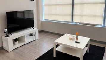 Uma sala de estar moderna com uma televisão de tela plana em um suporte branco, uma pequena mesa branca com um controle remoto e um tapete preto.