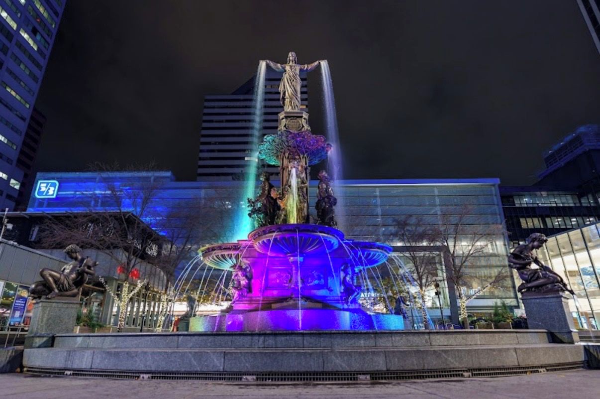 A nighttime view of a fountain with water jets and statues, surrounded by modern buildings.