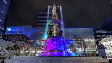 A nighttime view of a fountain with water jets and statues, surrounded by modern buildings.