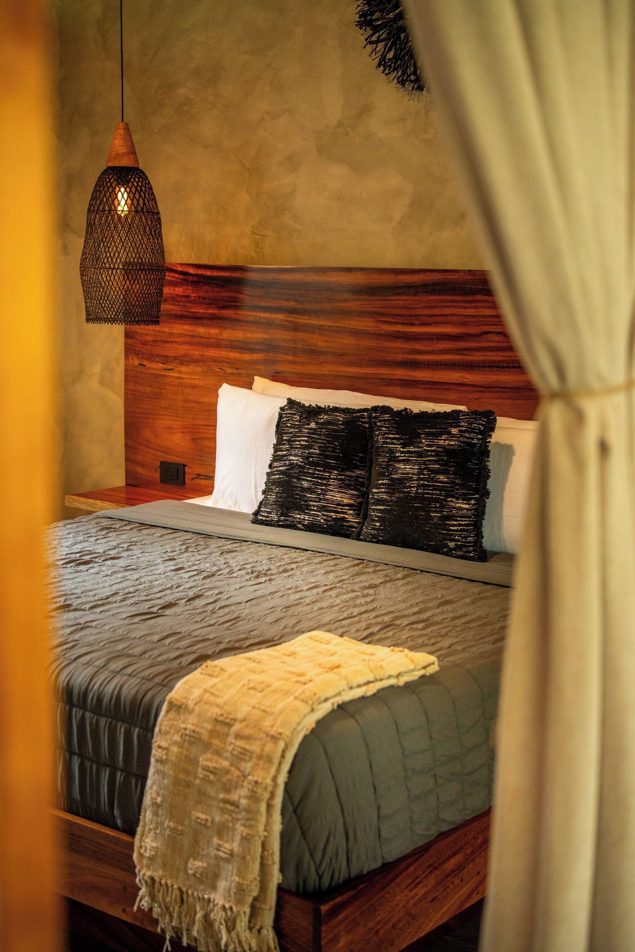 Cama com cabeceira de madeira, luminária pendente e manta texturizada.