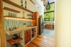 Une cuisine moderne avec des étagères en bois, un réfrigérateur encastré et une vue sur l’extérieur grâce à de grandes fenêtres.