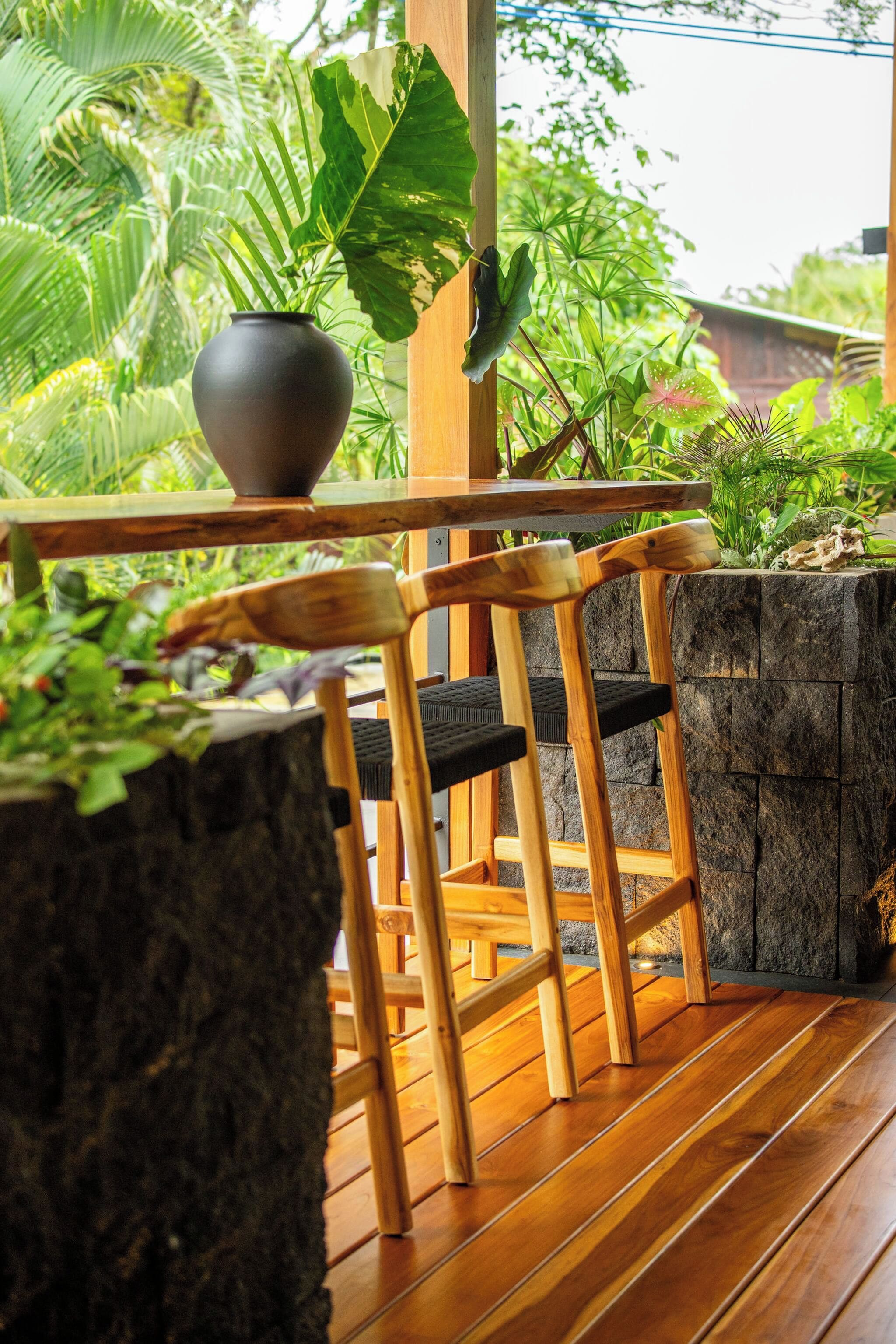 Uma área de estar ao ar livre com bancos de madeira, uma mesa, plantas em vasos grandes e uma parede de pedra.