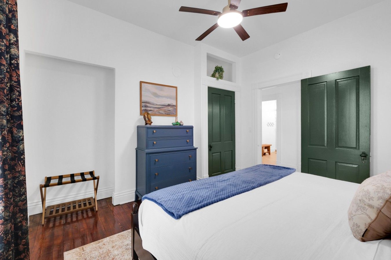 A bedroom with a bed, a dresser, two doors, a ceiling fan, and a painting on the wall.