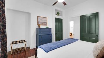 A bedroom with a bed, a dresser, two doors, a ceiling fan, and a painting on the wall.