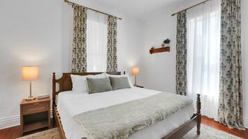 Ein Schlafzimmer mit einem hölzernen Bett, zwei Nachttischen inklusive Lampen, einer Zimmerpflanze und Vorhängen mit Blättermotiv.