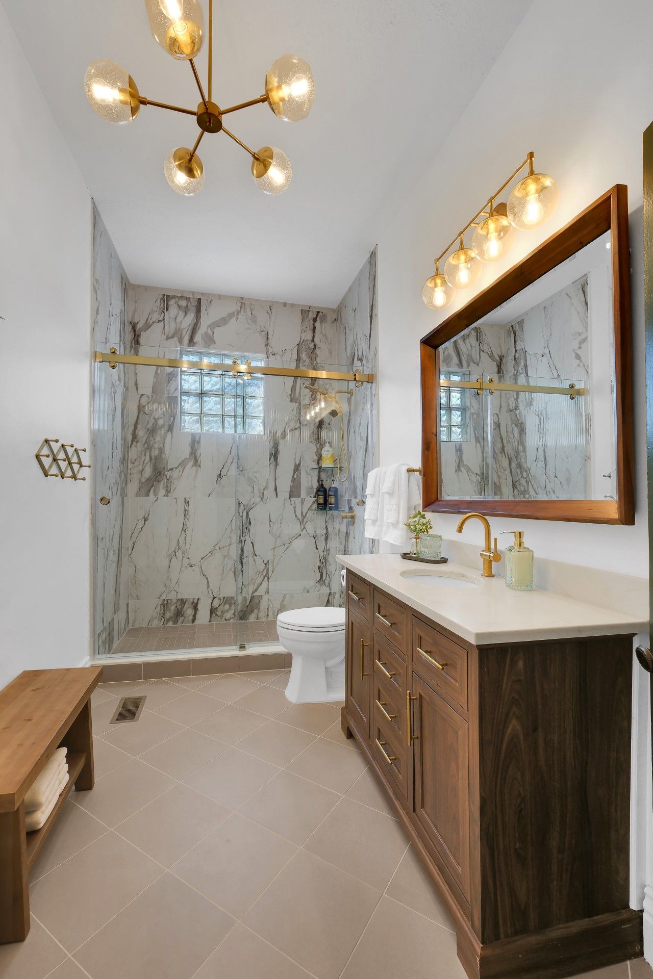 Un baño con pared de mármol, ducha, inodoro, tocador con espejo y un banco.