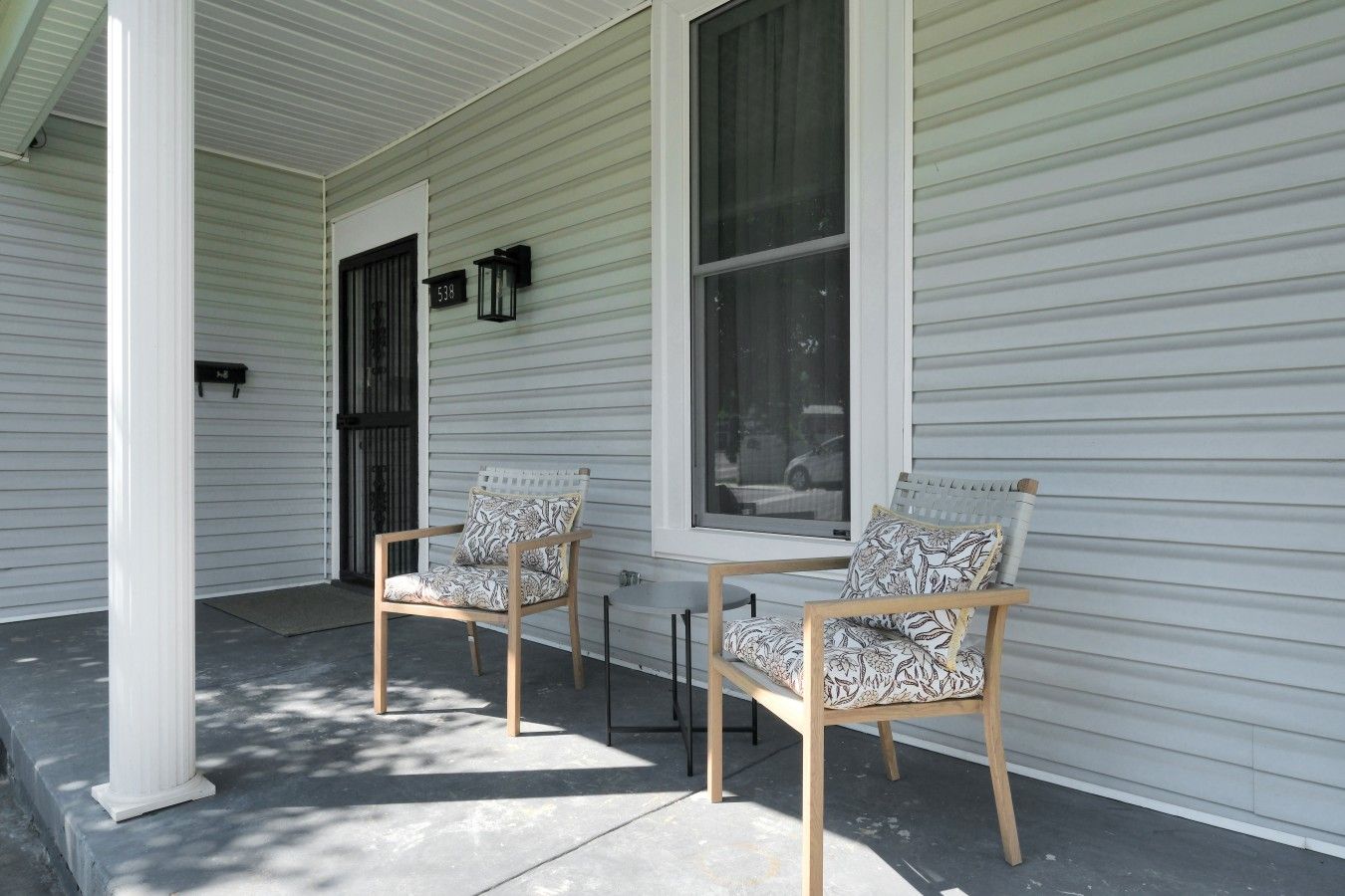 A porch with two wooden chairs and a table, white siding, and a window.