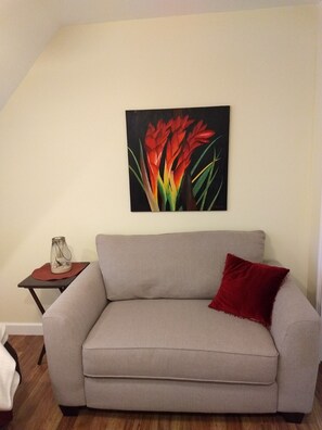 En stue med en beige sofa, en rød pude, en glassvase på et lille bord og et maleri af røde blomster på væggen.