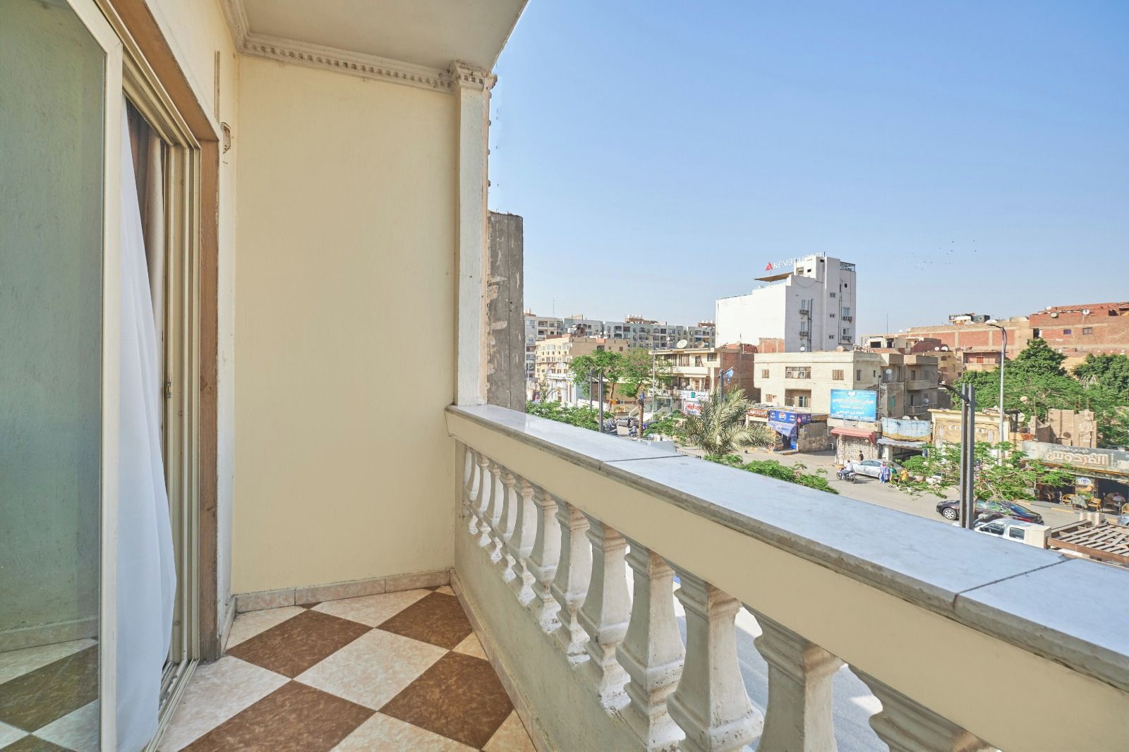 A balcony with a checkered floor, overlooking a cityscape with buildings and a clear sky.