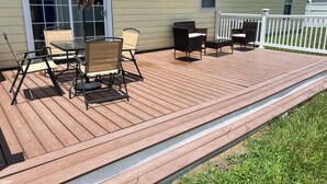 Een houten terras met buitenmeubilair, een overdekte tafel en een witte omheining.