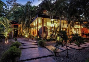 Una casa moderna amb façana de vidre, envoltada de plantes tropicals i un camí de pedra.