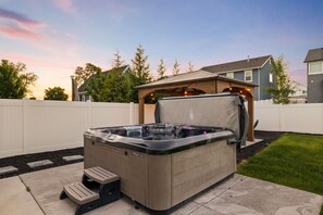 Een hottub met een houten terras en een pergola, gelegen in een achtertuin met een witte schutting en huizen op de achtergrond.