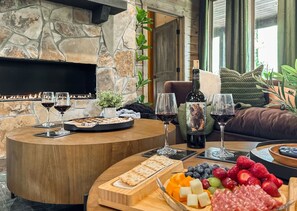 Um ambiente acolhedor com lareira de pedra, mesa redonda de madeira com petiscos e taças de vinho, e uma área de estar confortável com almofadas.