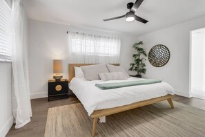 Ein Schlafzimmer mit Bett, Nachttisch, Lampe, Deckenventilator, Fenster mit Vorhängen, Zimmerpflanze und Wanddekoration.