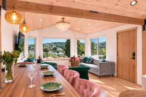 Amplia habitación con techo de madera, ventanas grandes que ofrecen vistas a una ladera, zona de comedor con una mesa larga y un área de estar con sofá y sillones.