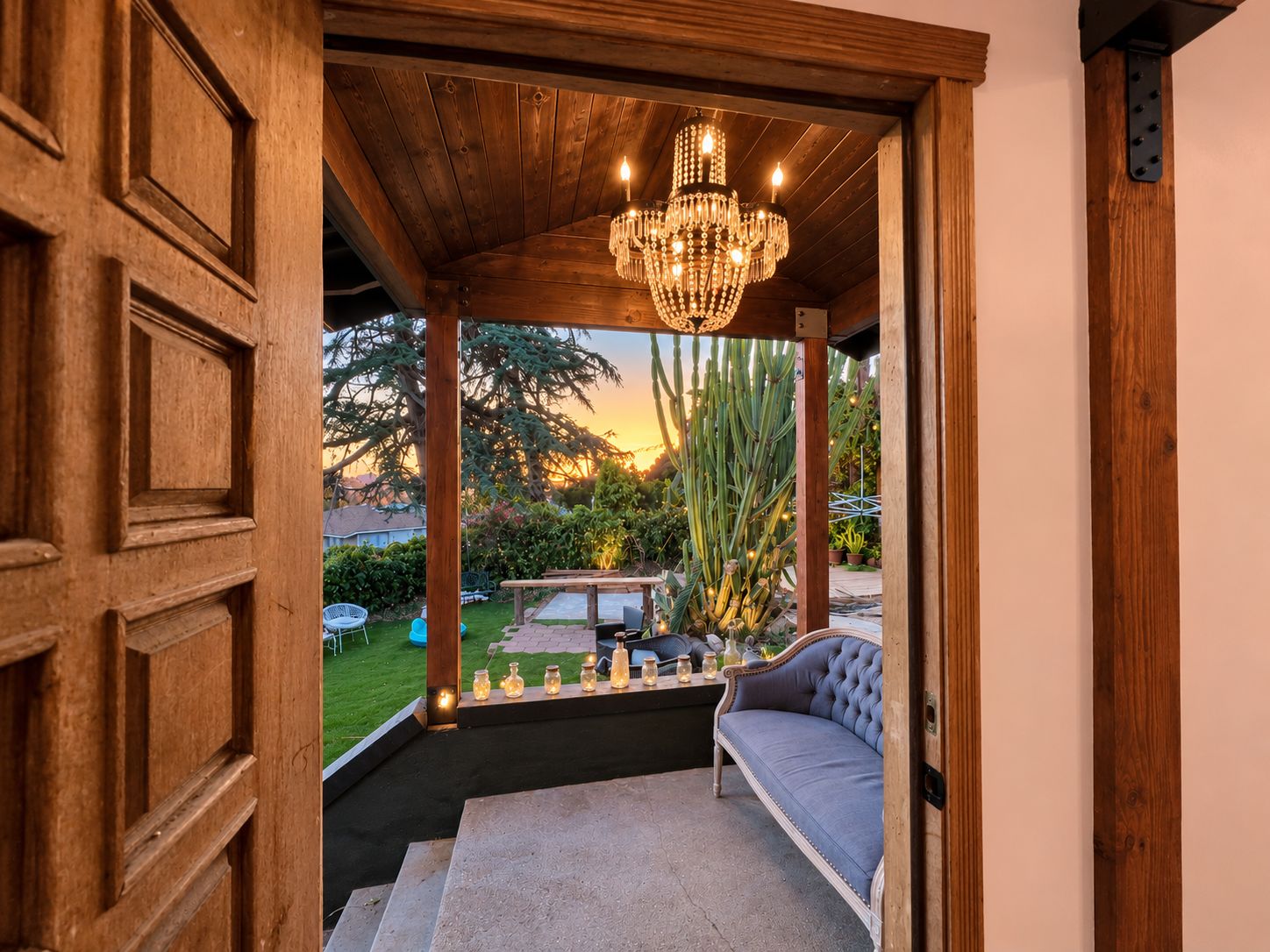 Un intérieur en bois avec un banc, un lustre et une vue sur un jardin agrémenté de cactus et illuminé par le coucher du soleil.