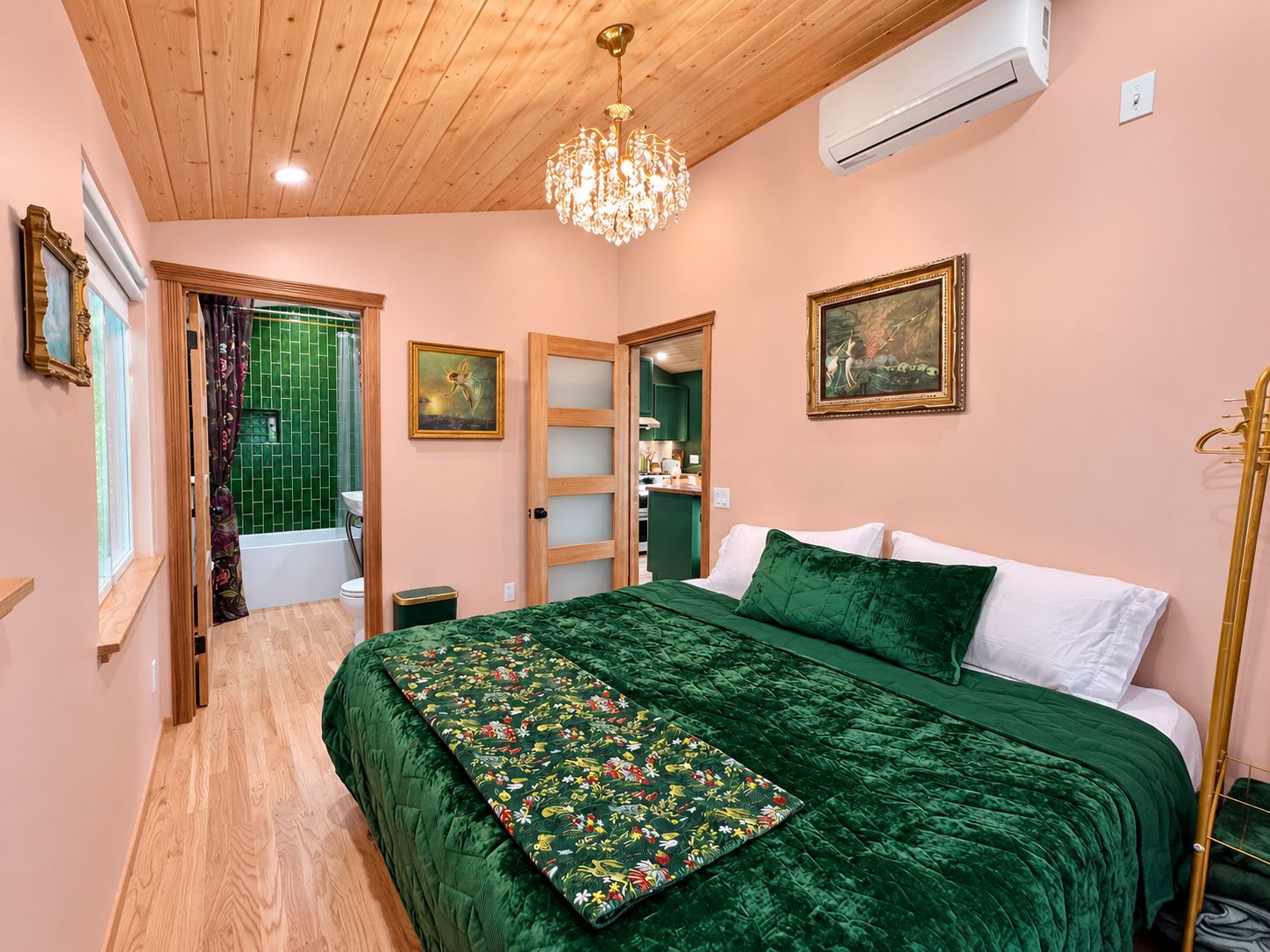 A bedroom with a wooden ceiling, a chandelier, a bed with green bedding, a green floral throw, and a painting on the wall.