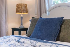 Ein Bett mit einem blumigem Überwurf, einem weißen Kopfteil und einer Lampe mit einem gewebten Schirm auf einem Nachttisch.