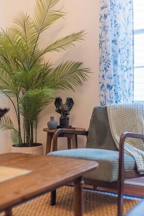 Ein Zimmer mit einem Holztisch, einem Sessel mit Kissen, einer Topfpflanze, einem kleinen Tisch mit einer Vase und einem Fenster mit Vorhängen.