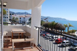 A balcony with a table and benches, overlooking a parking area and a coastal view with mountains in the distance. - Apartment in Nerja Near Carabeillo Beach (Nerja)