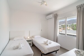 Ein Hotelzimmer mit zwei Einzelbetten, einem Nachttisch mit Lampe, einer Klimaanlage und einem Fenster mit Blick auf eine Stadt und Hügel.