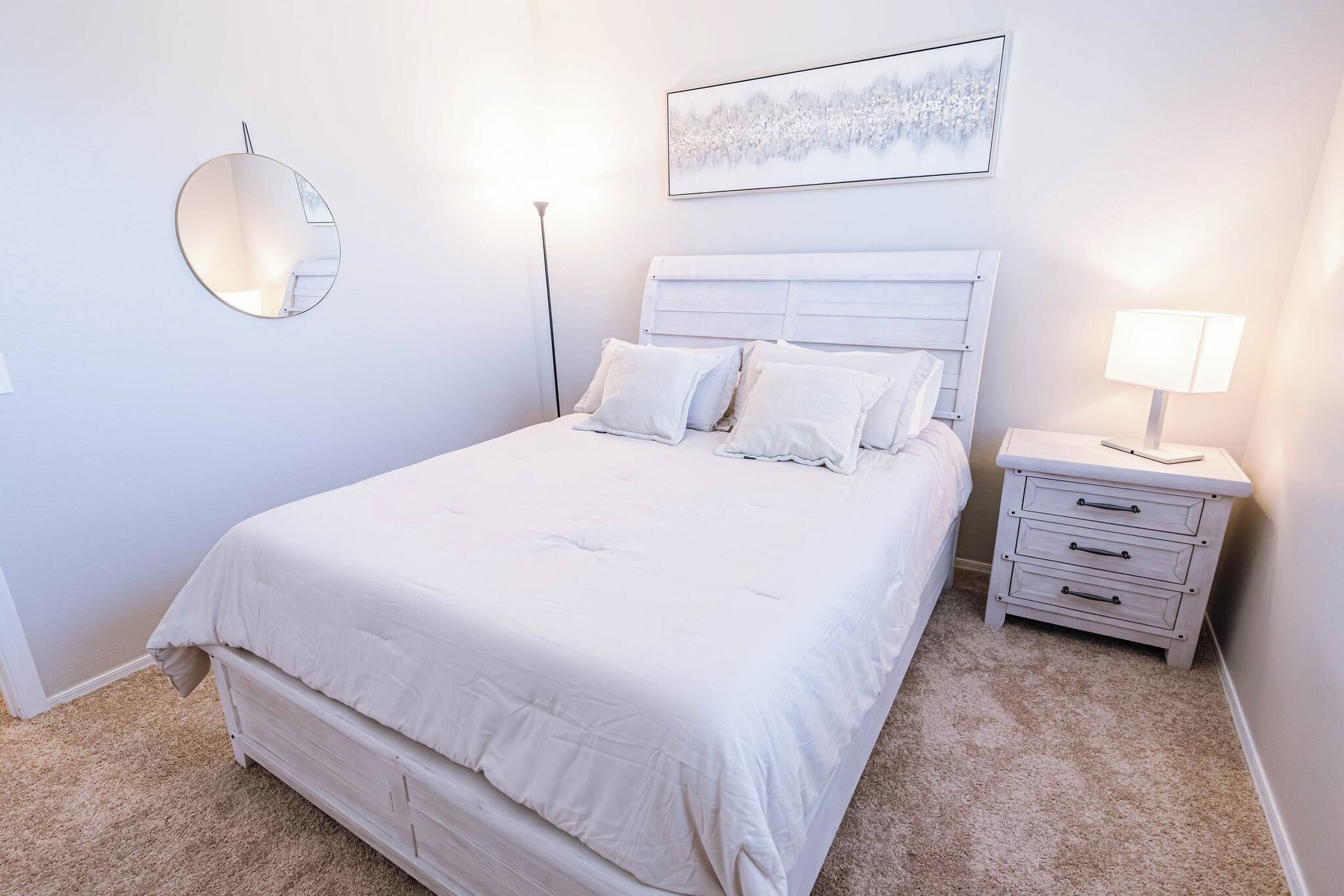 A bedroom with a bed, bedside tables, a nightstand, a lamp, a mirror, and a painting on the wall.
