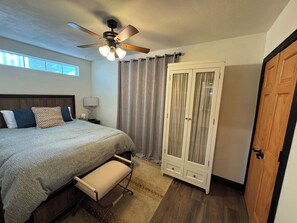 Une chambre à coucher comprenant un lit, une table de chevet, une commode, un ventilateur de plafond, une armoire et une porte.