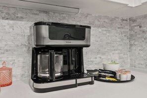 Máquina de café com jarra de vidro, bandeja com cápsulas de café e caneca branca em bancada.