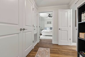 A hallway with white doors, a ceiling fan, a bed with a patterned bedspread, and a rug on the floor. - HiTide Condo | Walk to Campus | Prime Gameday Unit (Tuscaloosa)