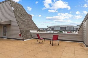 Een terras op het dak met twee rode stoelen, een brandblusser en een uitzicht op een complex van gebouwen in de achtergrond.