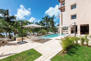 A pool area with lounge chairs and umbrellas, surrounded by palm trees and a building with balconies. - Artila Isla Mujeres: The Ocean Dream Sanctuary (Isla Mujeres)