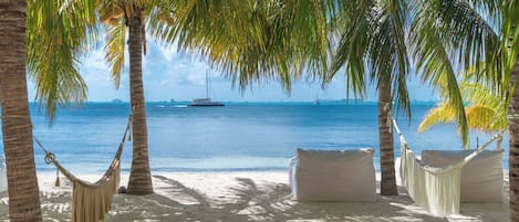 A beach with a hammock and white cushions under palm trees, overlooking the sea.