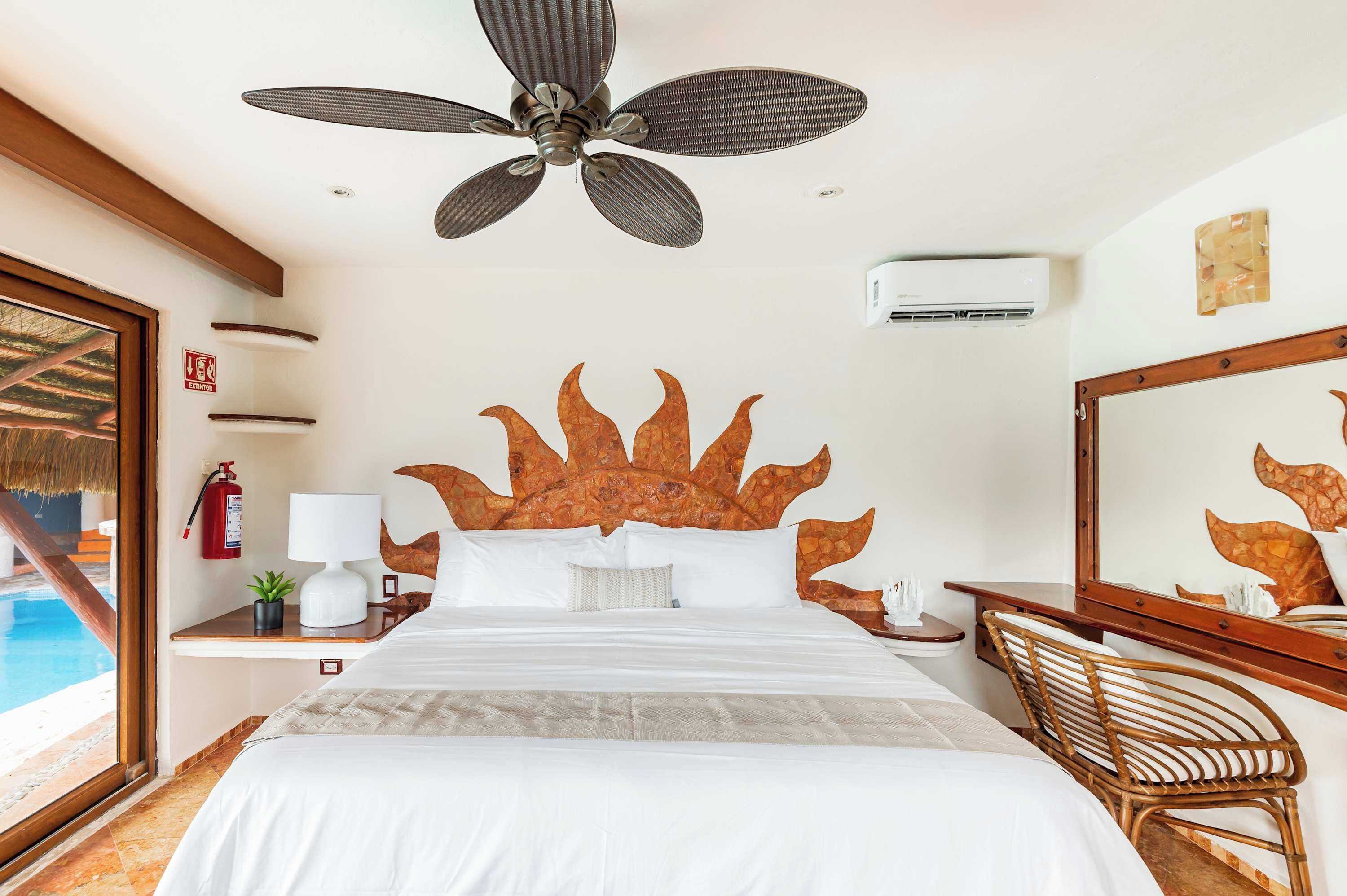 A hotel room with a bed, a ceiling fan, a nightstand, a chair, a large mirror, and a view of a swimming pool.