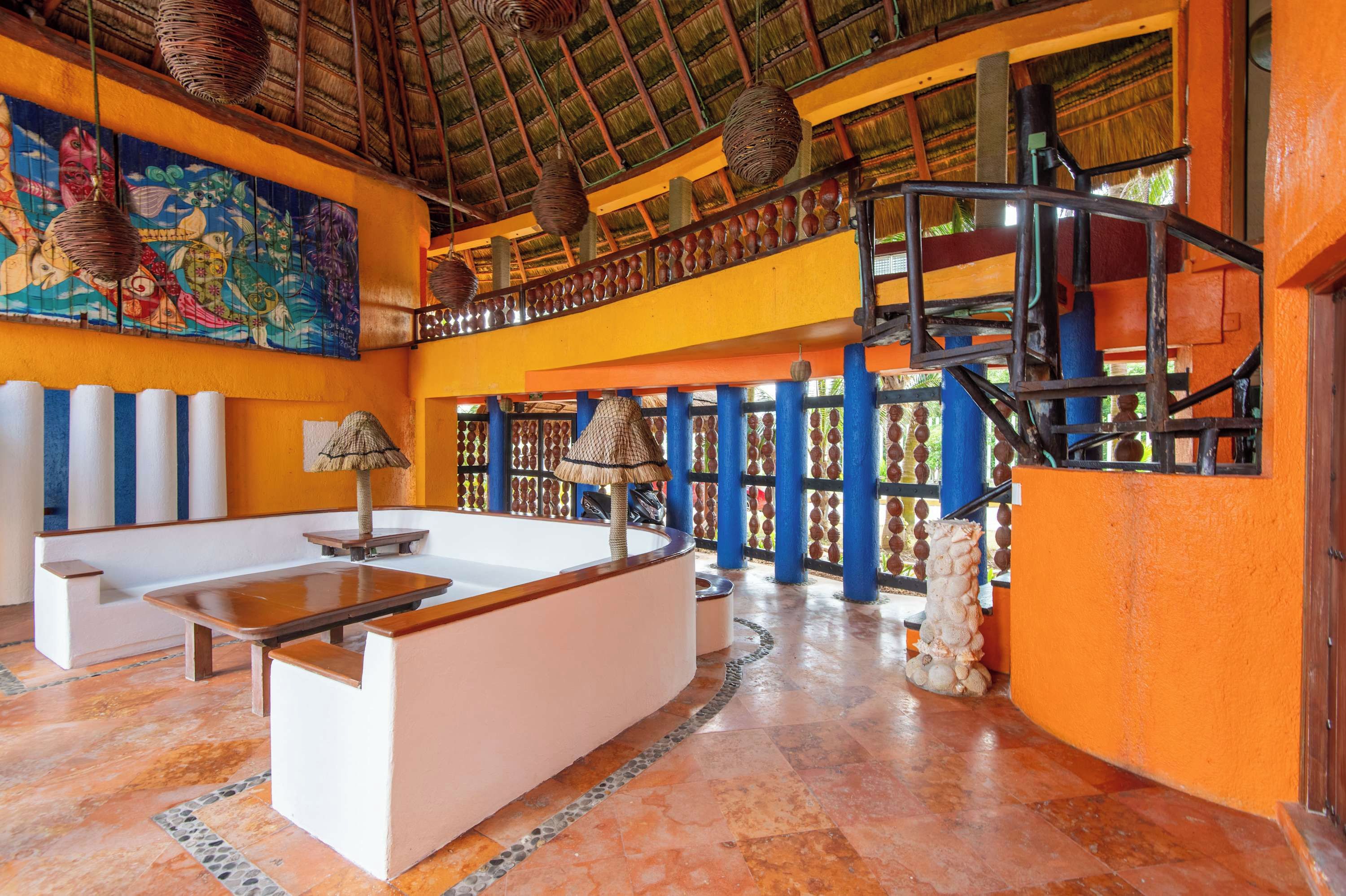 Um hall de hotel com teto alto, telhado de palha, um mural colorido, um balcão de recepção e uma escadaria.