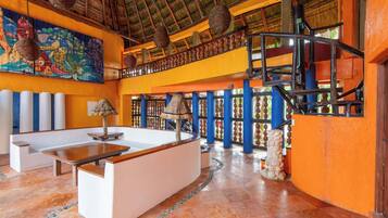 Um hall de hotel com teto alto, telhado de palha, um mural colorido, um balcão de recepção e uma escadaria.