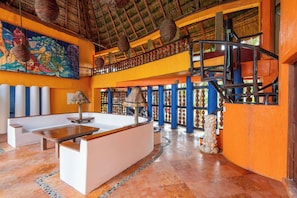 A hotel lobby with a high ceiling, a thatched roof, a colorful mural, a reception desk, and a staircase. - Bella Mar: Ocean Front w/ Pool Views (Isla Mujeres)