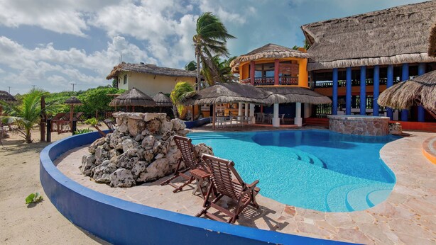 Uma área de piscina com espreguiçadeiras, um elemento decorativo de rocha e edifícios com telhados de palha.
