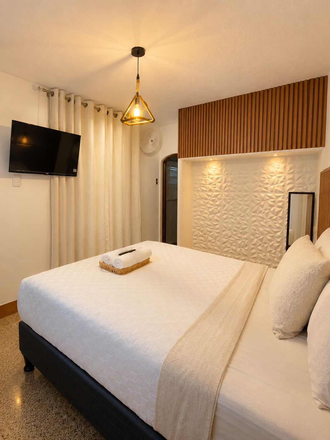 A hotel room with a bed, a flat-screen TV, a wall-mounted light fixture, and a textured wall panel.