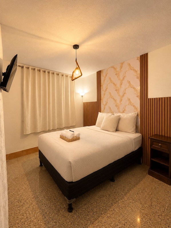 A hotel room with a bed, a flat-screen TV mounted on the wall, a bedside table, and a wall-mounted lamp.