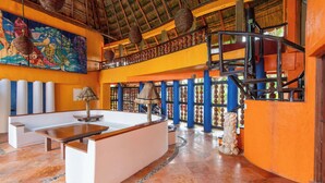 Um hall de hotel com teto alto, telhado de palha, um mural colorido, um balcão de recepção e uma escadaria.