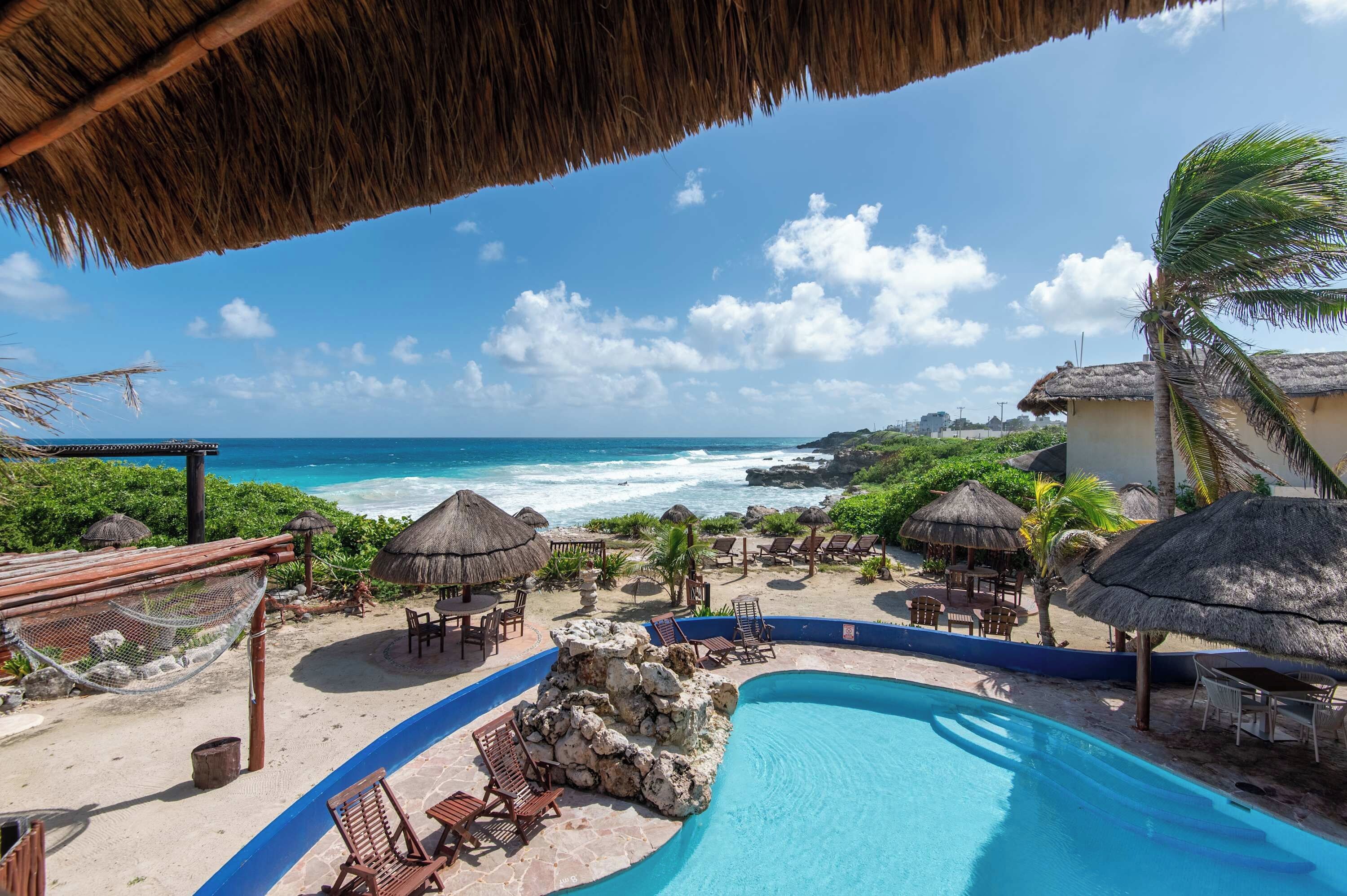 Ein Poolbereich in einem Resort mit Blick auf das Meer, mit Reetdachschirmen und Liegestühlen.