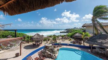 Ein Poolbereich in einem Resort mit Blick auf das Meer, mit Reetdachschirmen und Liegestühlen.