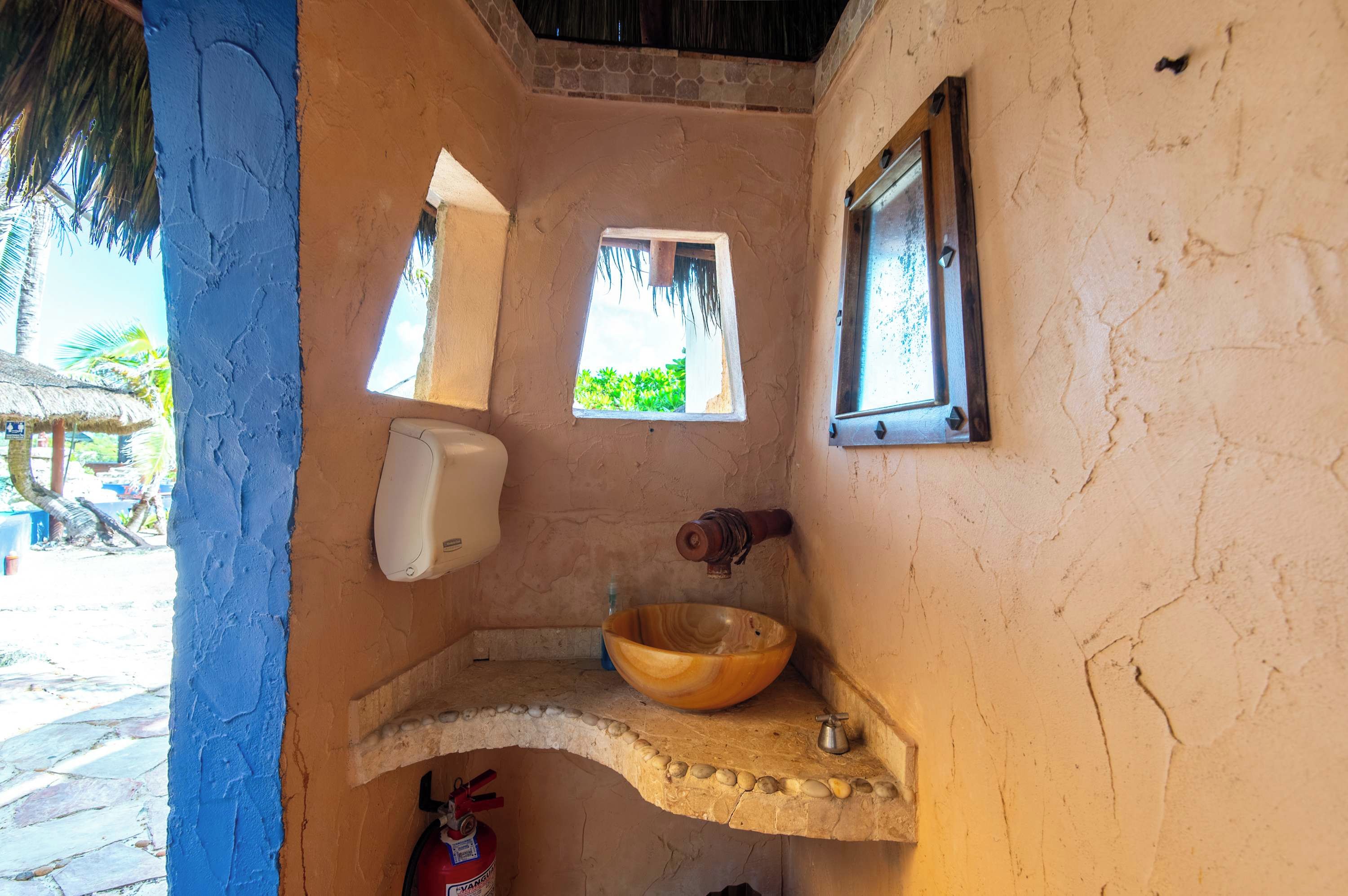 Casa de banho pequena com lavatório de pedra, uma tigela de madeira, dispensador de sabonete líquido e janela com vista para o verde.