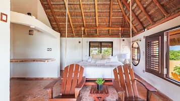 Un dormitorio con una cama, sillas de madera, una mesita, una planta en maceta y una ventana con contraventanas.