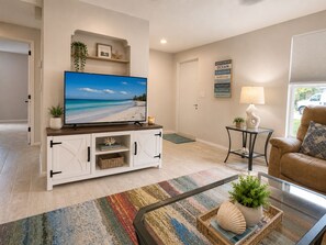Ein Wohnzimmer mit einem Flachbildfernseher auf einer weißen Kommode, einem braunen Ledersofa, einem Glastisch und einem farbenfrohen Teppich.